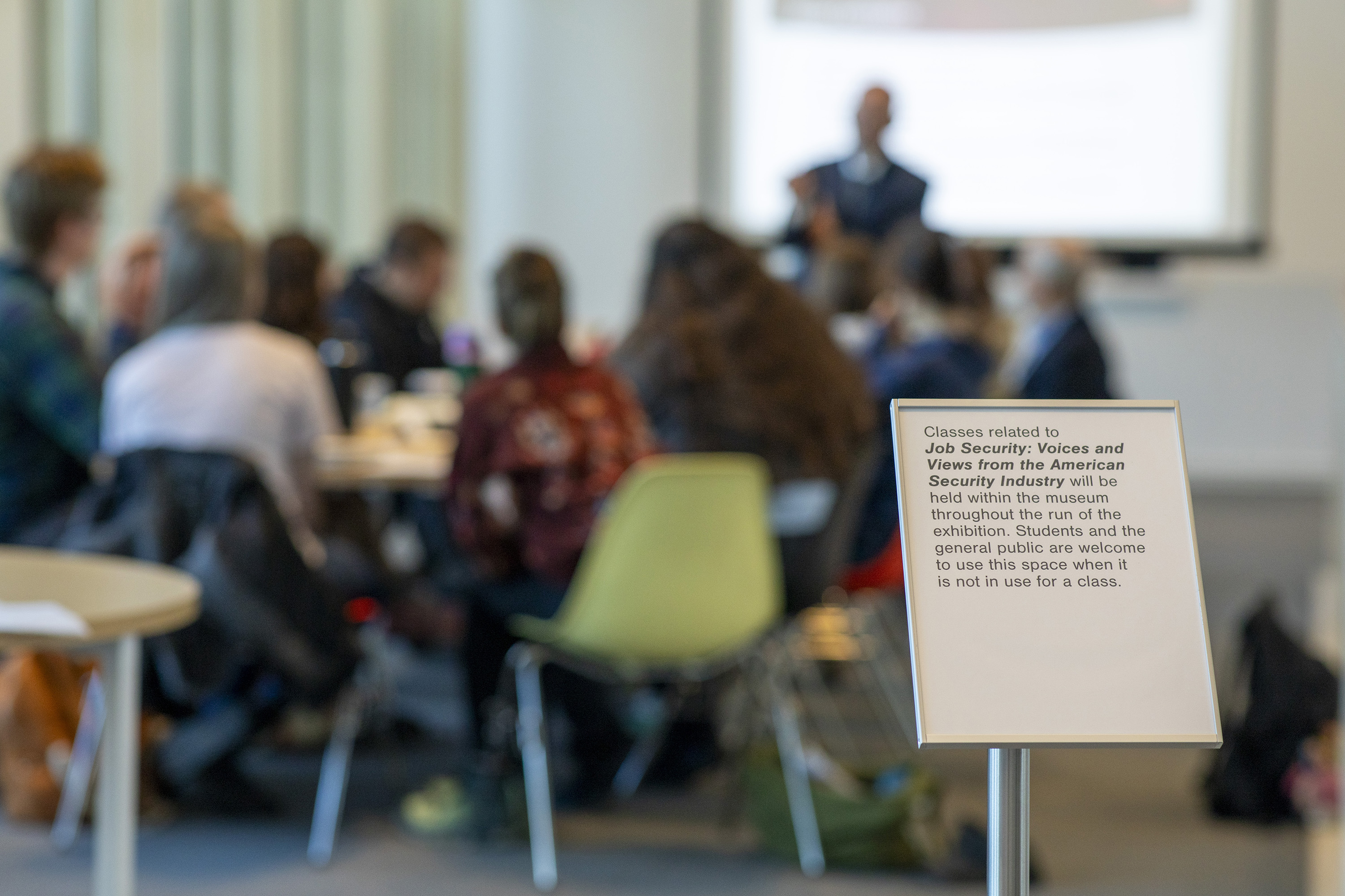 Shallow focus photograph of a presenter in front of a class with an aluminum sign in the foreground. Sign reads: Classes related to Job Security: Voices and Views from the American Security Industry will be held within the museum throughout the run of the exhibition. Students and the general public are welcome to use this space when it is not in use for a class.